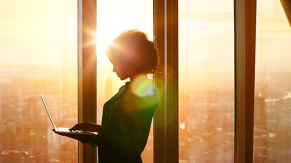 Ženska, ki ob sončnem zahodu stoji za okni v stavbi in uporablja prenosnik