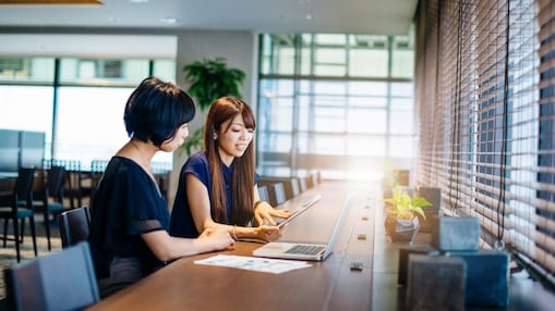 Dve ženski sedita za mizo in gledata v računalnik in tablico