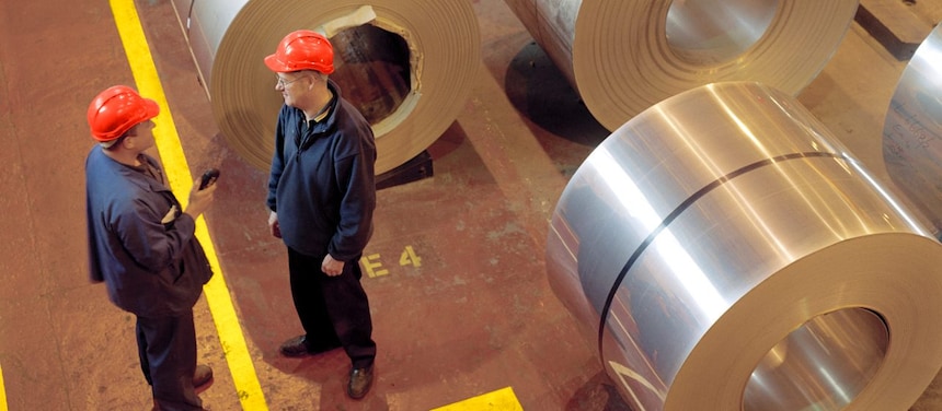 Two factory workers talking next to large rolls of steel
