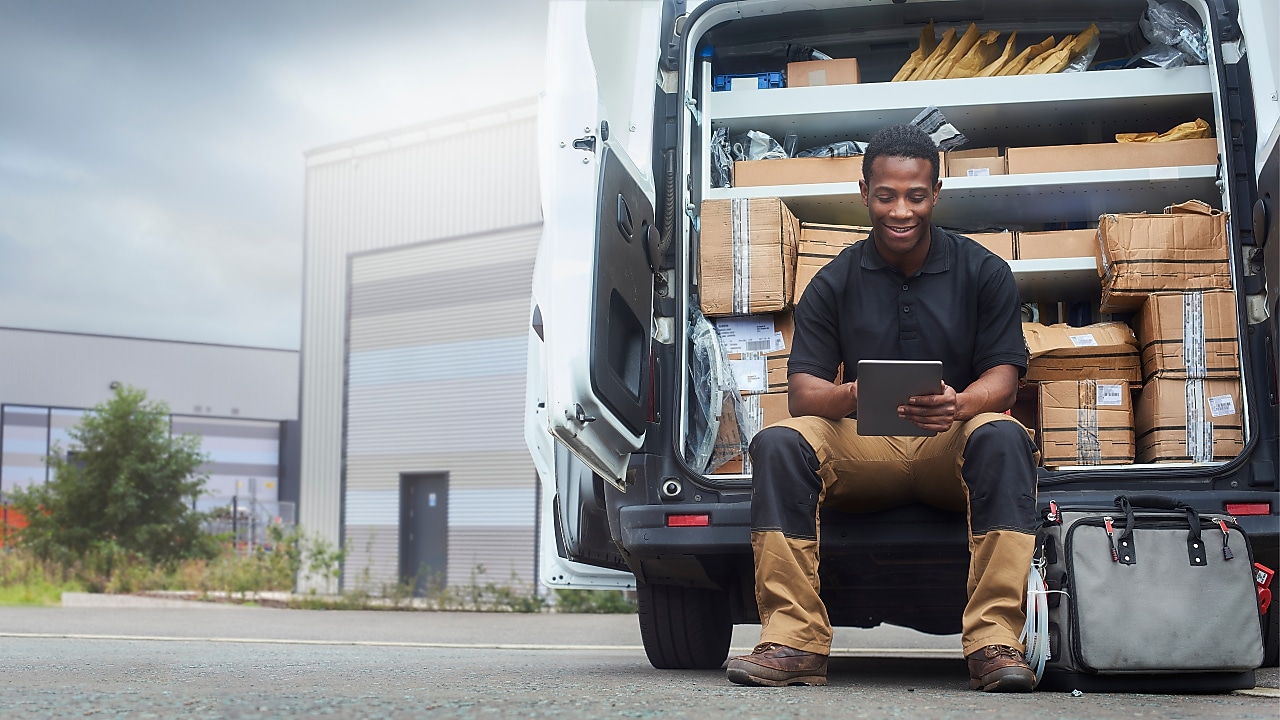 Dostavljavec sedi na robu odprtega dostavnega kombija, obdan s paketi in škatlami, ter uporablja tablični računalnik. V ozadju je industrijska stavba, na tleh pa torba z orodjem.