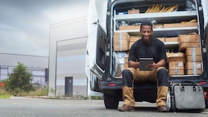 Dostavljavec sedi na robu odprtega dostavnega kombija, obdan s paketi in škatlami, ter uporablja tablični računalnik. V ozadju je industrijska stavba, na tleh pa torba z orodjem.