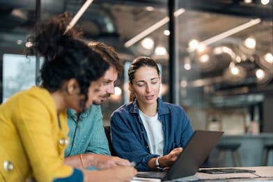 Small group of co-workers working together in a startup business office space. A team of young people in a modern office is discussing their project. Group of business people working. The team are sitting at a board room table looking at a laptop computer.