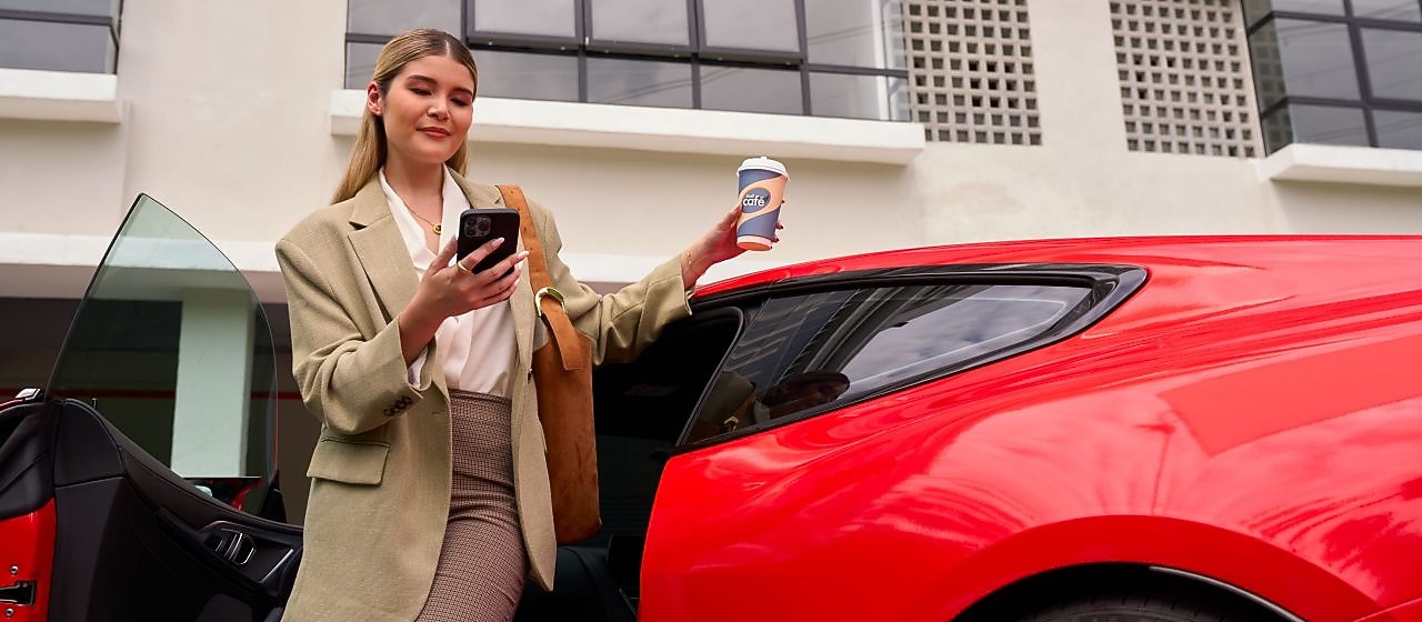 Poslovna ženska izstopa iz rdečega avtomobila, gleda v telefon in drži skodelico Shell Café.