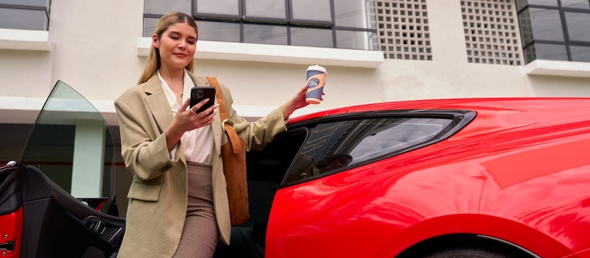 Poslovna ženska izstopa iz rdečega avtomobila, gleda v telefon in drži skodelico Shell Café.